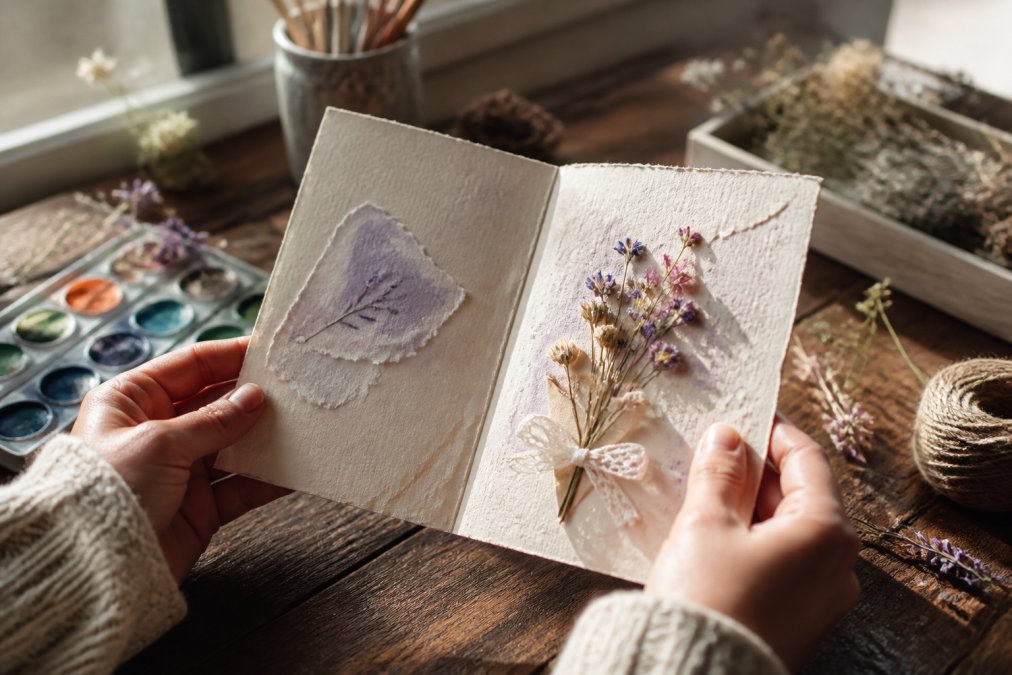 Close-up of hands holding an open handmade greeting card with visible texture, embellishments, and hand-painted details caught in natural window lighting