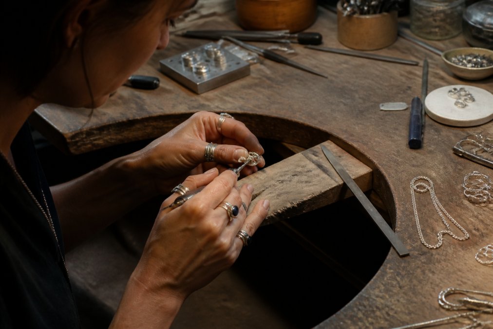 Close-up of a silversmith's hands carefully working on a piece of jewelry at a workbench, with tools, materials, and finished pieces visible
