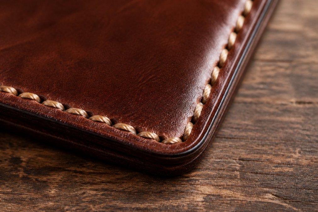 Close-up of hand-stitched saddle stitch seam on leather wallet showing wax-coated thread and individual stitches with burnished leather edges, natural lighting highlighting craftsmanship detail