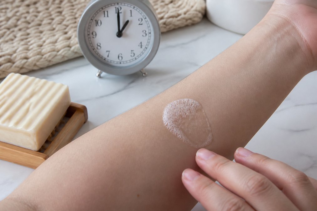 Close-up of a person's forearm with a small patch of lather from a handmade soap being gently tested on the inner arm skin, with a timer visible in the background showing the 24-hour observation period