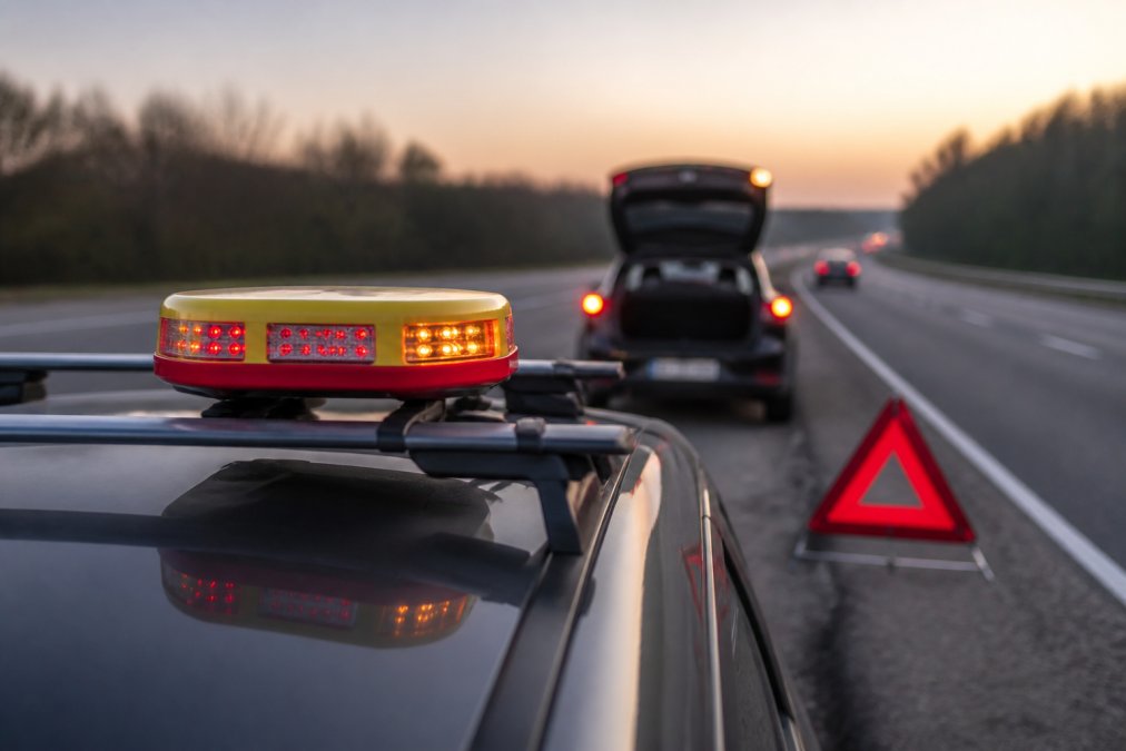 Why Hazard Lights Are Not Enough Close-up of a GPS-enabled emergency beacon mounted on a vehicle's roof rack with high-visibility reflective surfaces and LED indicators, positioned next to a traditional reflective warning triangle on a motorway hard shoulder at dusk