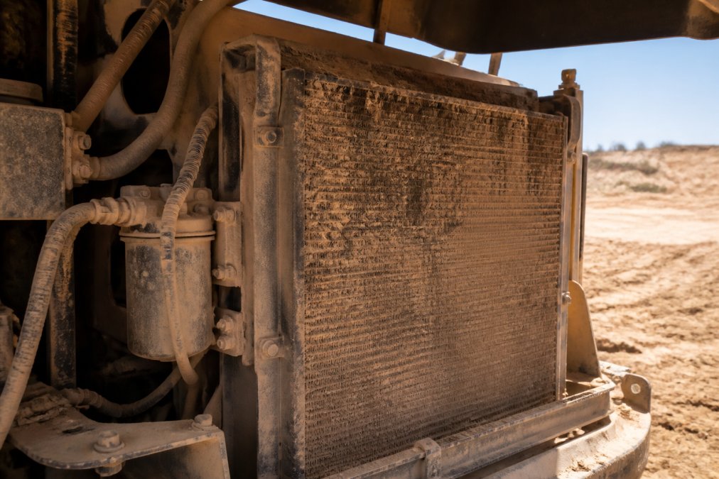 Close-up of a dusty, clogged condenser coil on an earthmoving machine in a dry, sunlit construction site, with visible dust buildup between the fins