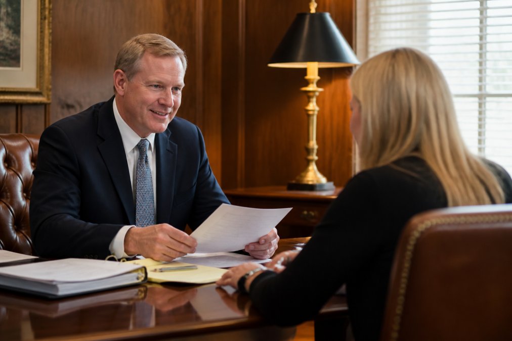 A professional attorney in a business suit sitting across a desk from a client, reviewing documents together in a warm, wood-paneled office with natural light