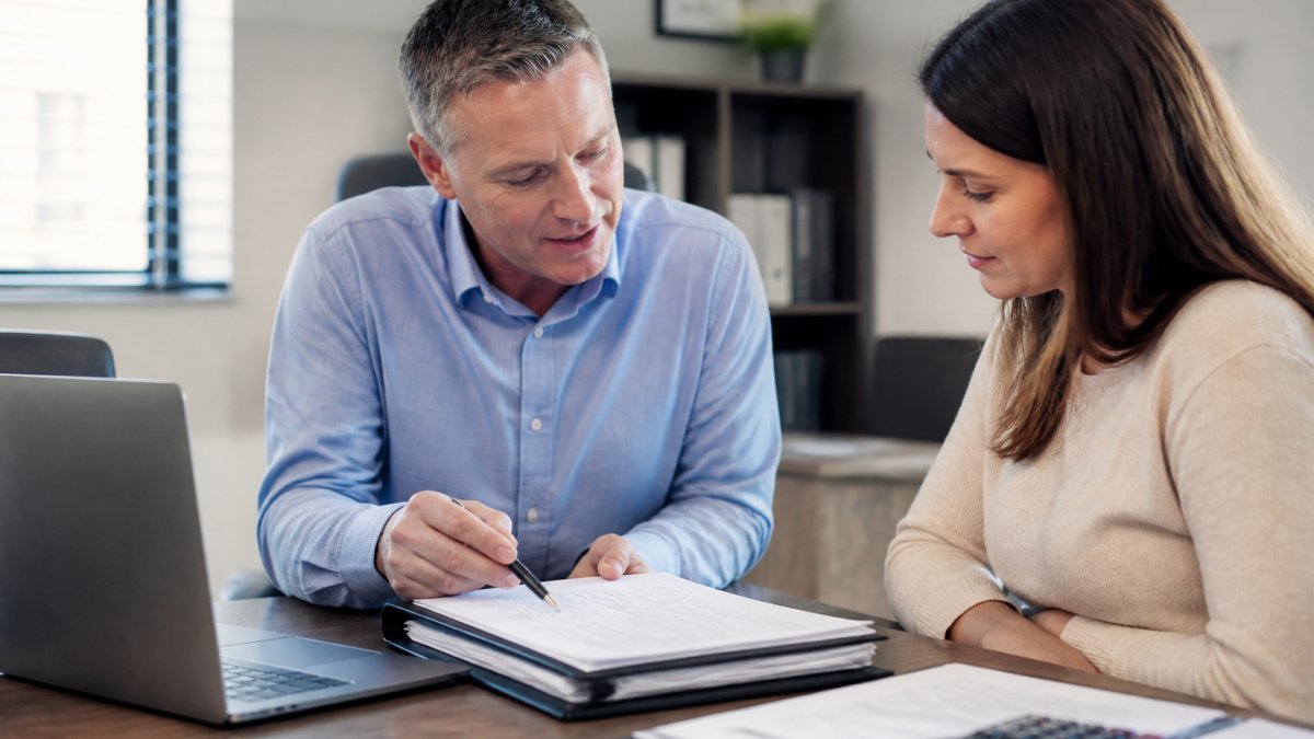 A tax professional and a client reviewing documents together at a desk in a bright office, with a laptop and a folder of tax paperwork visible