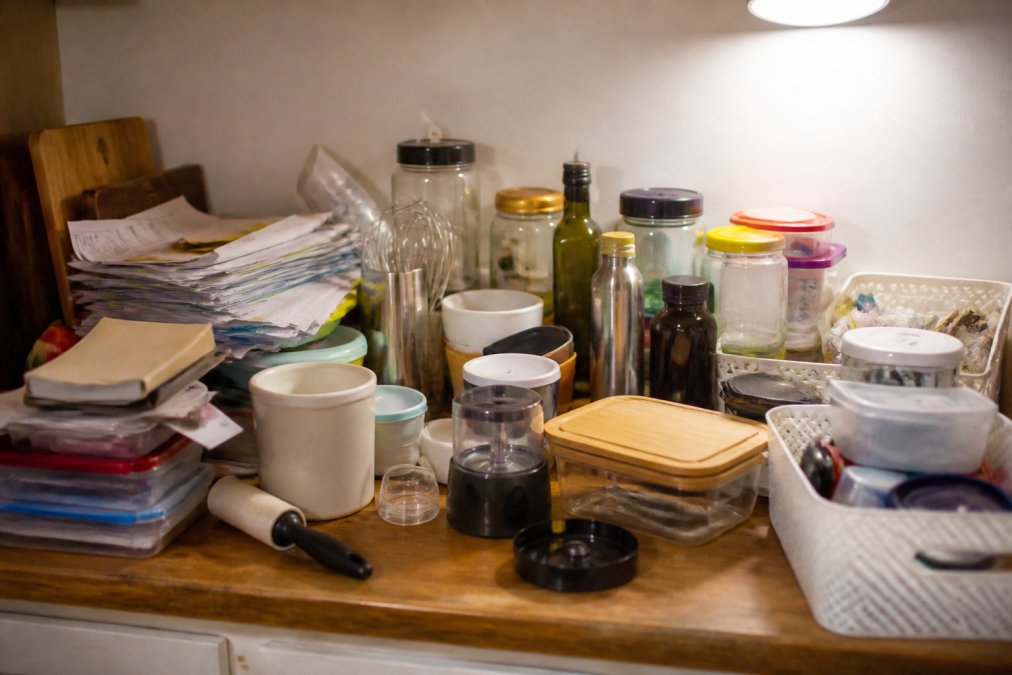 A cluttered kitchen counter covered with mismatched containers, unused gadgets, and stacked papers spread across a worn wooden surface under harsh overhead lighting, showing the chaos before a decluttering session begins
