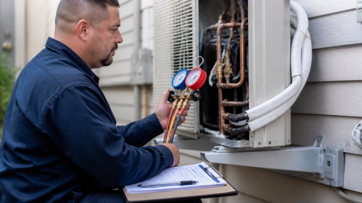 Professional HVAC technician in uniform examining a residential air conditioning unit mounted on exterior wall, holding diagnostic tools and clipboard, focused on inspecting copper refrigerant lines