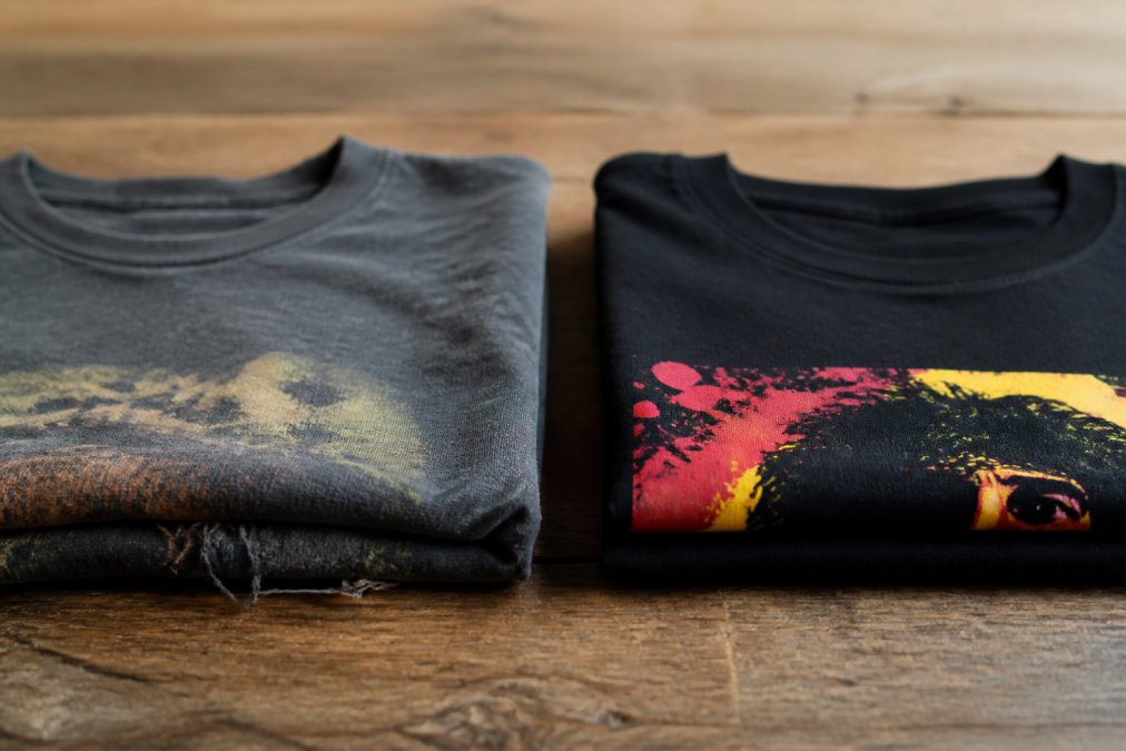 Close-up comparison of two folded t-shirts on a wooden table: one thin, faded mass-produced shirt with loose threads next to a thicker, well-constructed premium tee with crisp print