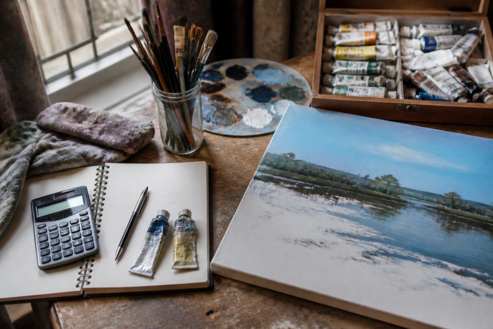 A close-up overhead shot of a professional artist's studio desk, showing high-quality paint tubes, brushes, and a canvas in progress, with natural window light illuminating the scene