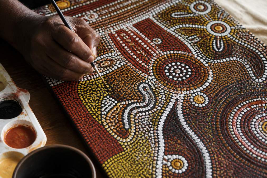 Close-up of traditional Indigenous artwork showing intricate dot patterns, layered brushwork, and symbolic designs in ochre and natural pigments on canvas