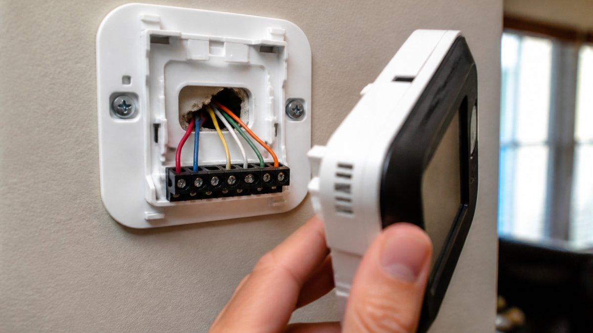 Close-up of a smart thermostat being pulled off its wall base plate, exposing the R, C, Y, W, G, and O/B terminal wires