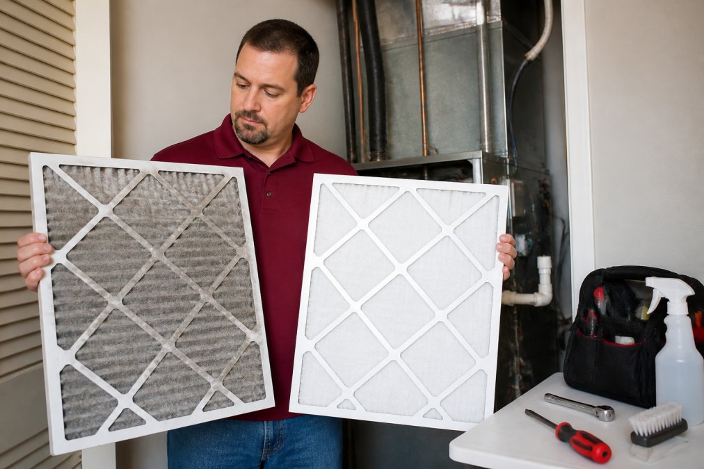 Close-up of a homeowner holding a visibly dirty gray HVAC air filter next to a clean white replacement filter, standing near an indoor air handler unit in a utility closet with overhead fluorescent lighting