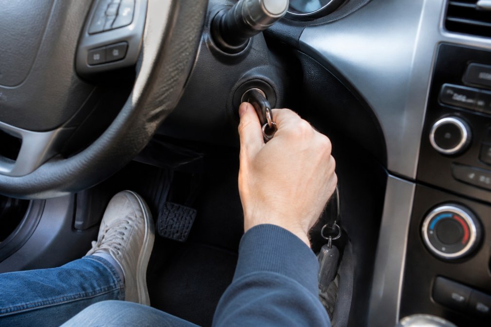 Close-up of a driver's hands gently wiggling a car key in the ignition while pressing the brake pedal, with the steering wheel visible in a modern car interior
