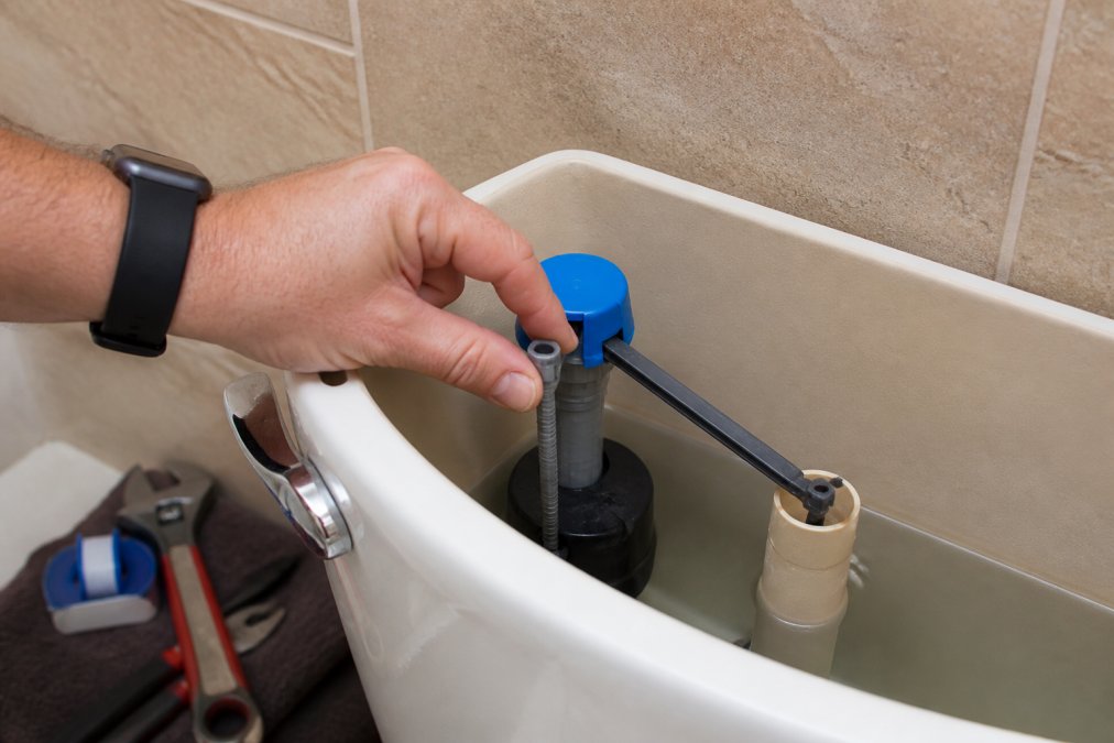 A person's hand adjusting the float cup on a vertical fill valve inside an open toilet tank, with the water level line and overflow tube clearly visible in the background, bathroom tiles in soft natural light