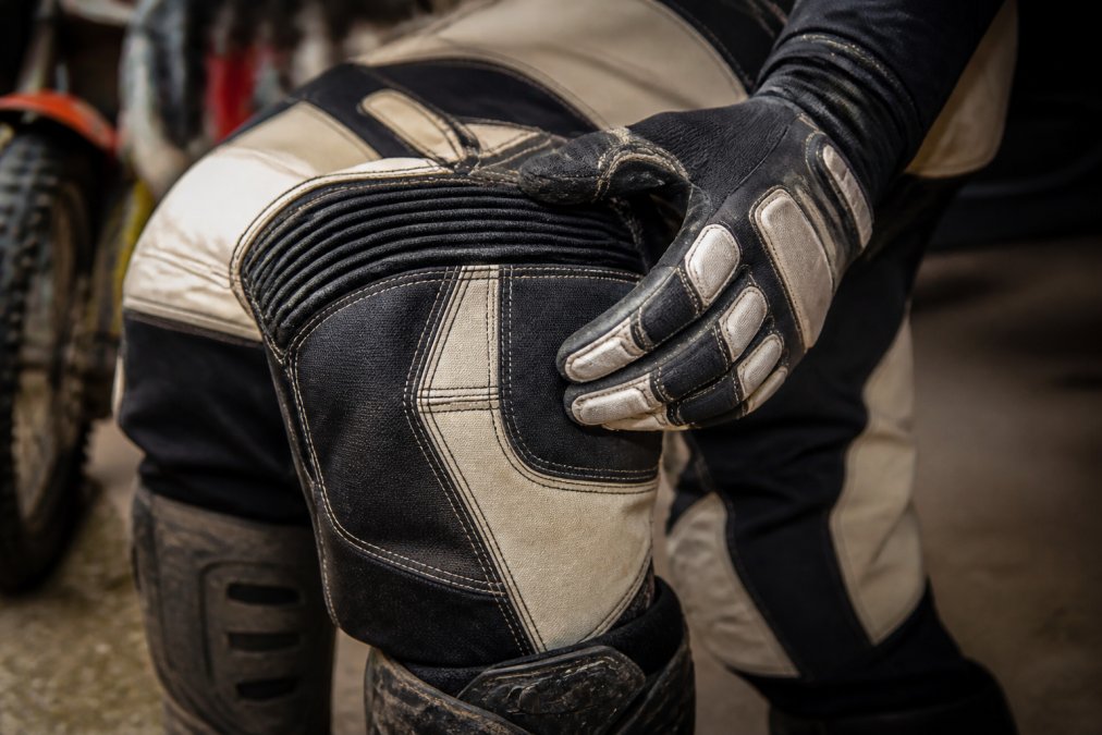 Close-up of a rider's gloved hand gripping the reinforced knee area of premium motocross pants, showing multi-layer panel construction and heavy-duty stitching in a dusty garage
