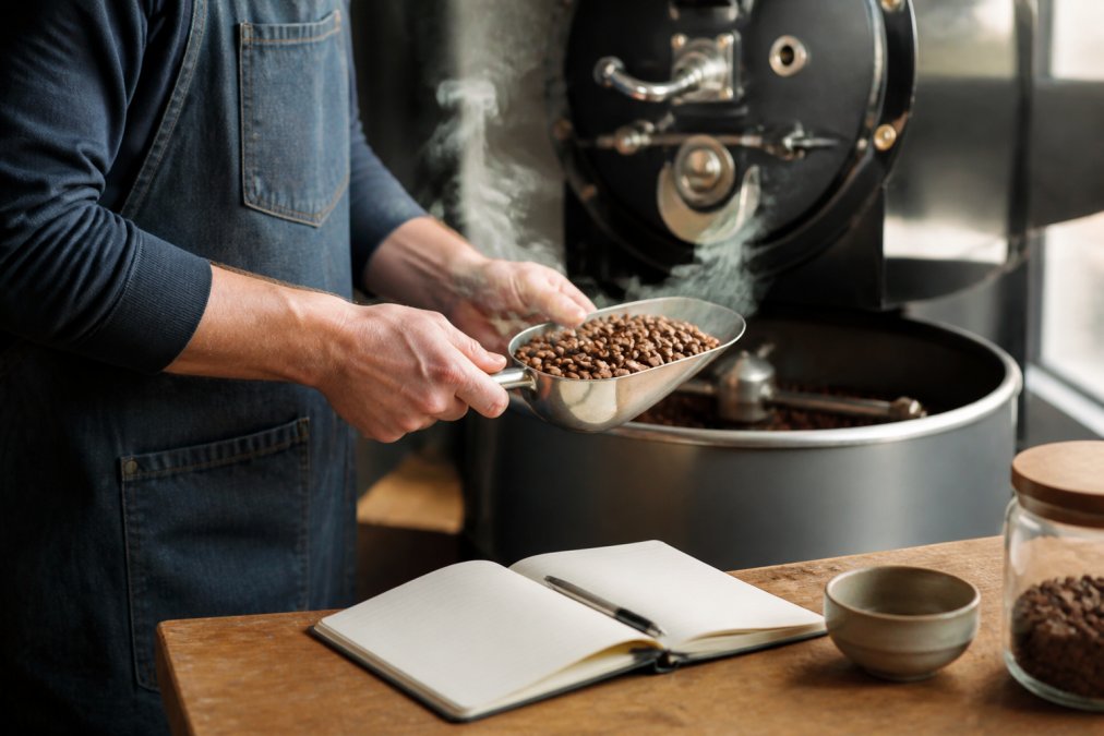 Close-up of a roastmaster's hands holding a metal scoop of freshly roasted coffee beans next to a small drum roaster, with steam rising and a notebook of roast notes visible on a wooden table
