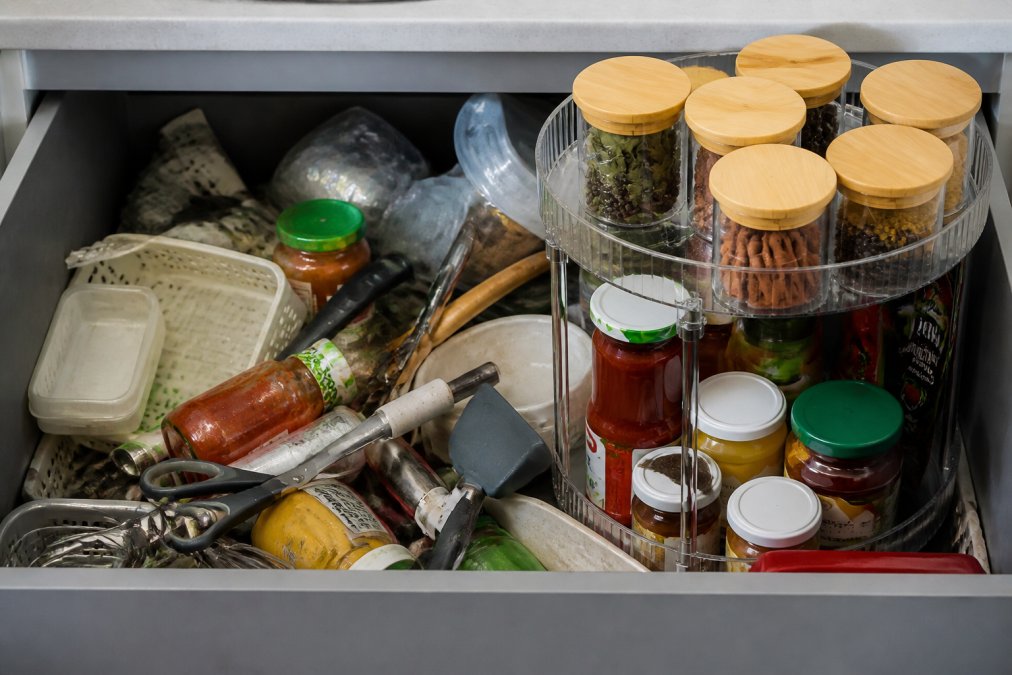 Cluttered kitchen drawer with mismatched organizers, expired items visible, and various kitchen tools stacked haphazardly showing visual chaos and disorganization