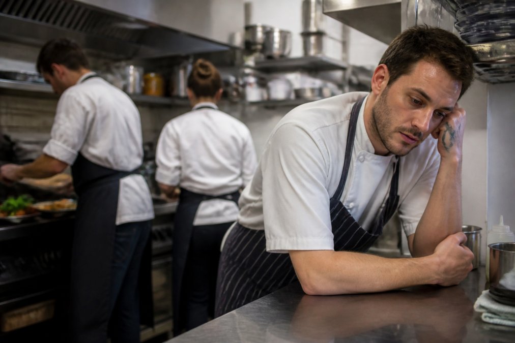 A tired chef in a busy commercial kitchen leaning against a stainless steel counter during a quiet moment, with other kitchen staff working in the background