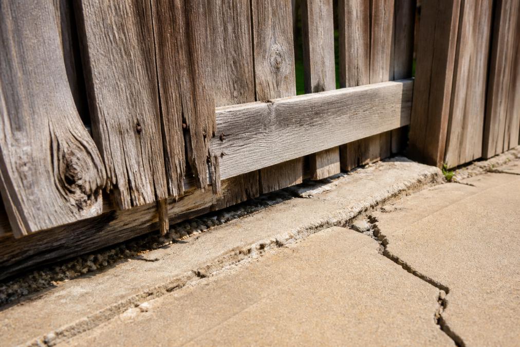 Close-up of warped, cracked wooden fence boards and concrete showing heat damage and weathering in bright sunlight