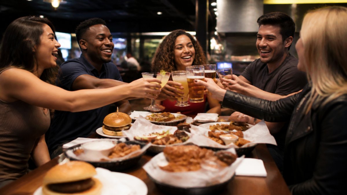 A diverse group of friends laughing and toasting around a table late at night, with plates of wings, burgers, and appetizers in the center, casual bar atmosphere with warm lighting