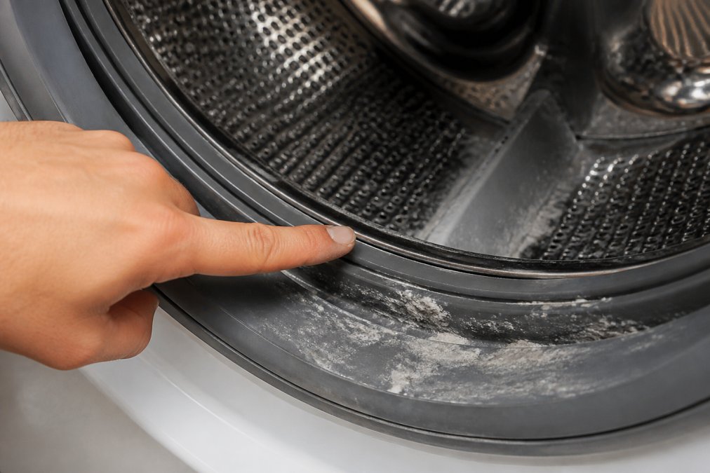 Close-up of a washing machine drum showing white detergent residue buildup on the rubber seal and drum ribs, with a hand pointing to the affected areas