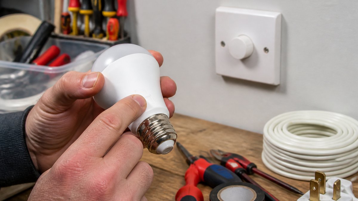 Close-up of hands examining the back of a dimmable LED bulb to check for the dimmable marking, with a dimmer switch visible in the background