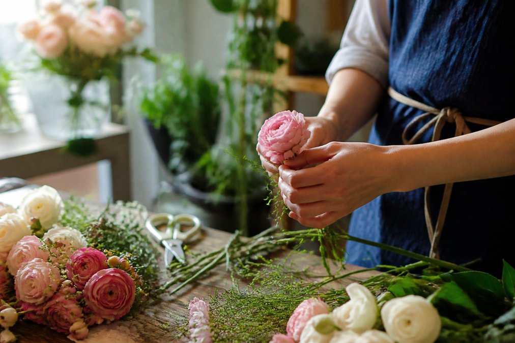 Florist hands carefully arranging fresh-cut flowers with delicate stems and vibrant petals in natural daylight, showing detailed precision work with scissors and floral materials