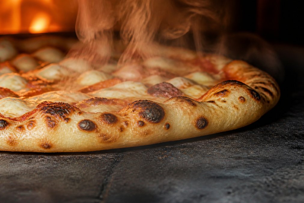 Close-up of a pizza crust with visible char bubbles and golden-brown spots, steam rising, on a wood-fired pie fresh from the oven