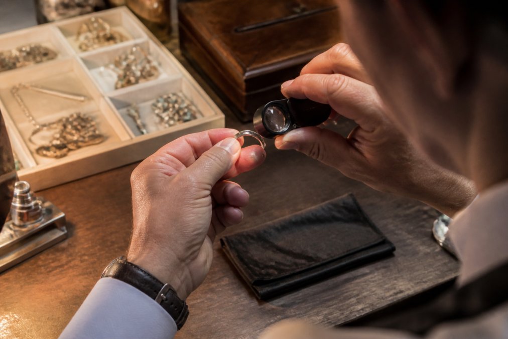 Close-up of a jeweler's hands holding a loupe to examine the inside of a silver ring, with a vintage jewelry tray and soft workshop lighting in the background