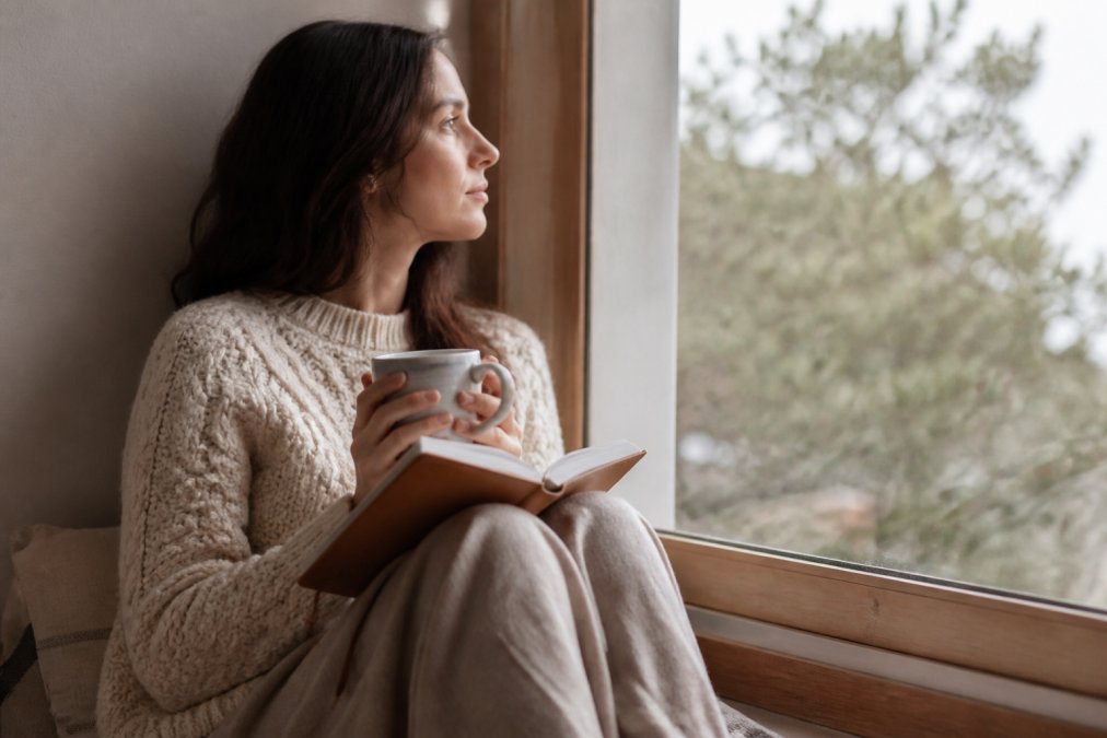A woman sitting quietly by a window with a journal and a cup of tea, looking thoughtful and calm, soft natural light coming through the window
