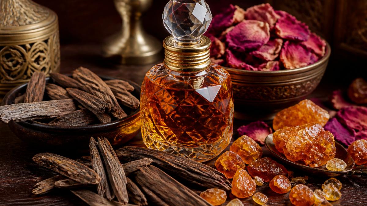 Close-up still life of an ornate glass perfume bottle surrounded by oud wood chips, dried rose petals, and amber resin on a dark wooden surface, warm side lighting