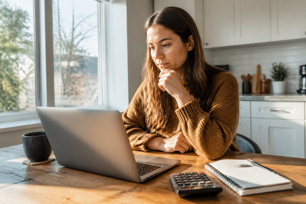 A homebuyer reviewing mortgage rate options on a laptop at a kitchen table, with a calculator and notepad nearby, morning light coming through a window