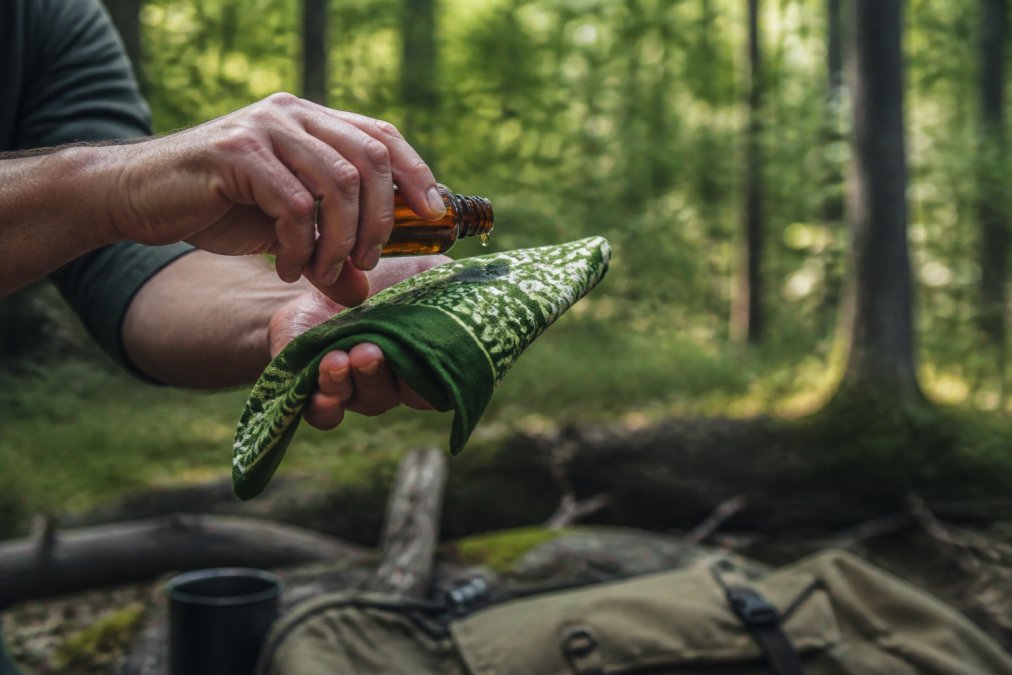 A close-up of a person applying a few drops of natural fly repellent from a small amber bottle onto a bandana while outdoors in a forest setting, with sunlight filtering through trees