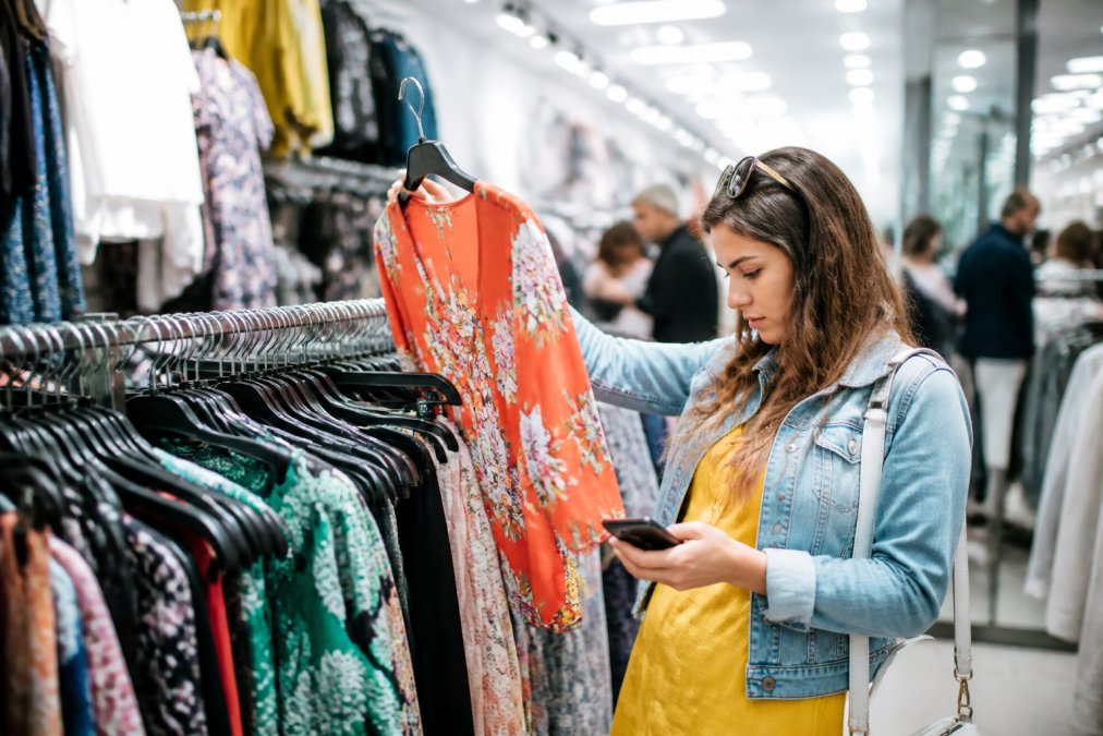 A woman browsing colorful clothing racks at a busy retail store, holding a garment up while checking her phone simultaneously under bright fluorescent lighting