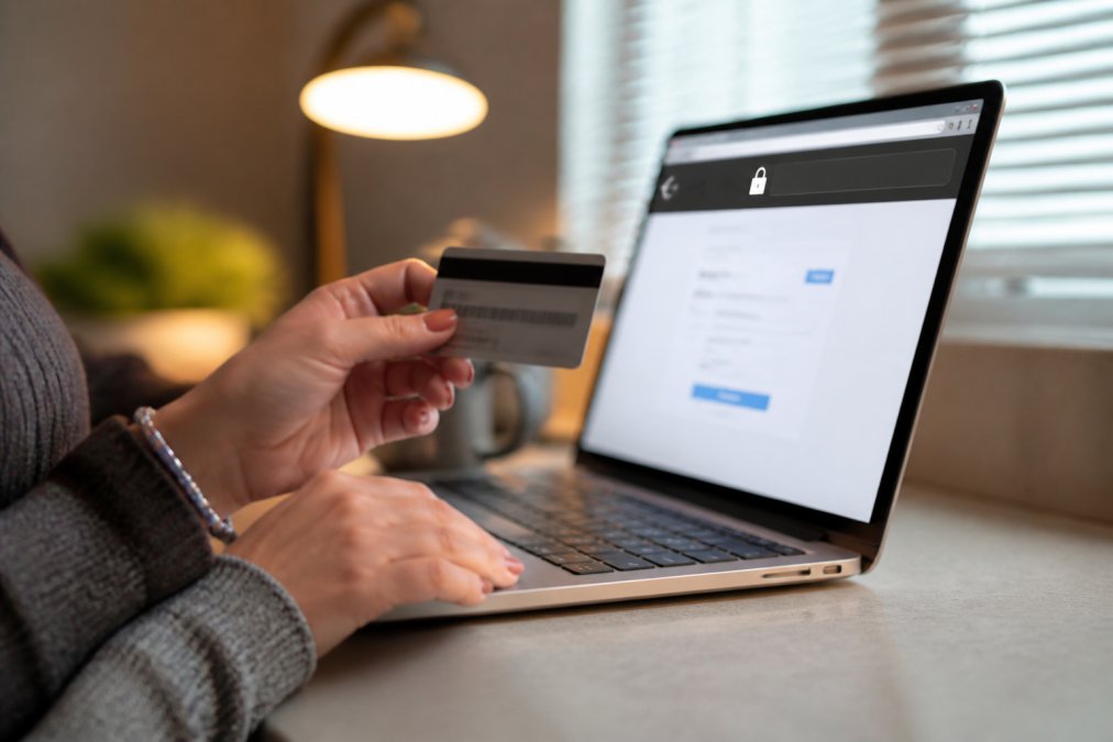 Close-up of a woman's hands holding a credit card near a laptop screen showing a secure checkout page with a padlock icon visible in the browser bar, warm desk lamp lighting