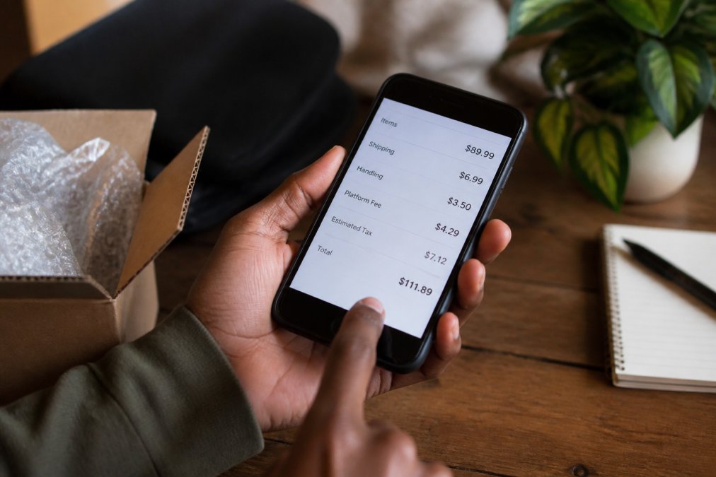 Close-up of a smartphone screen displaying a checkout page with itemized fees highlighted, including shipping, handling, and platform charges, with a shopper's hand pointing at the final total amount