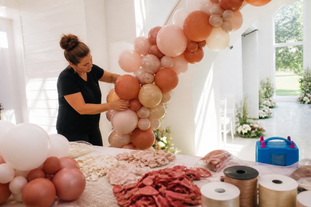 Professional balloon artist arranging asymmetrical clusters of balloons in warm earth tones and blush hues, creating depth and texture against a white wedding ceremony backdrop with natural light streaming across the installation