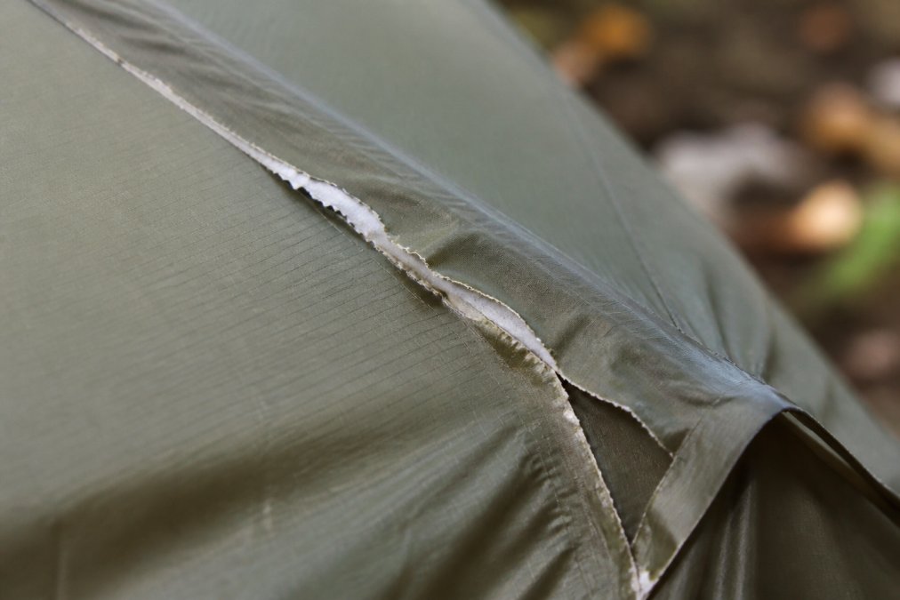 Close-up of a tent seam with visible tape peeling away from the fabric, showing white delamination and fraying edges under natural outdoor light