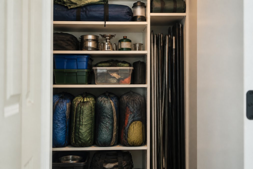 Interior of a well-organized storage closet with camping gear neatly arranged on shelves, sleeping bags in breathable mesh storage bags, and tent poles stored vertically in a dark space