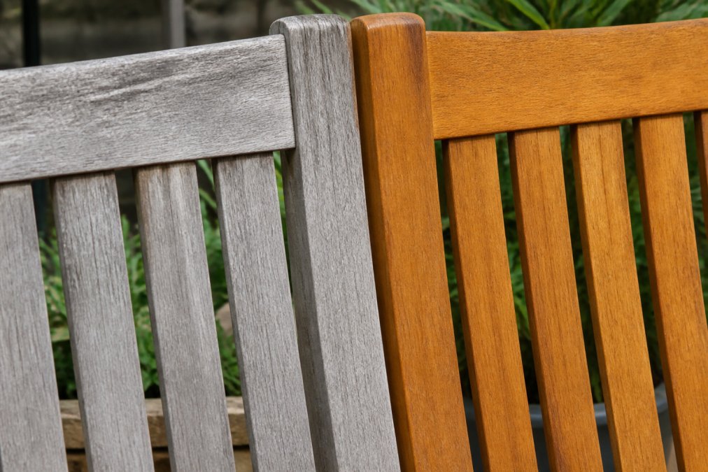 Close-up of weathered gray wood grain on an outdoor teak patio chair next to a section of freshly oiled golden-brown wood, showing the contrast between weathered and protected surfaces in natural sunlight