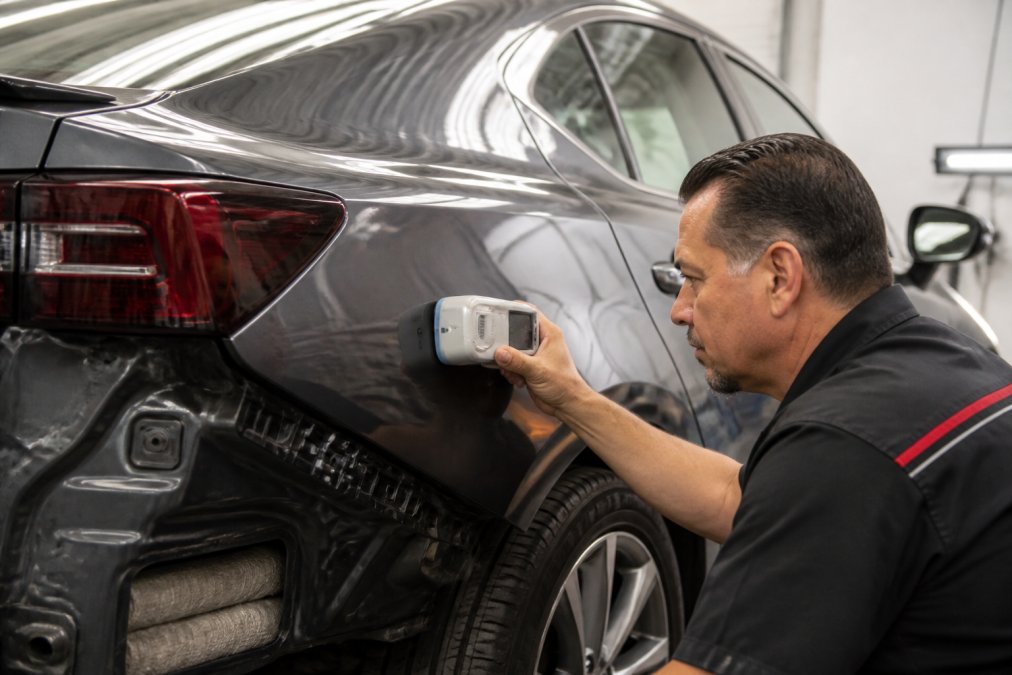 Professional auto body technician using a spectrophotometer device to measure and match paint color on a vehicle panel in a well-lit shop environment