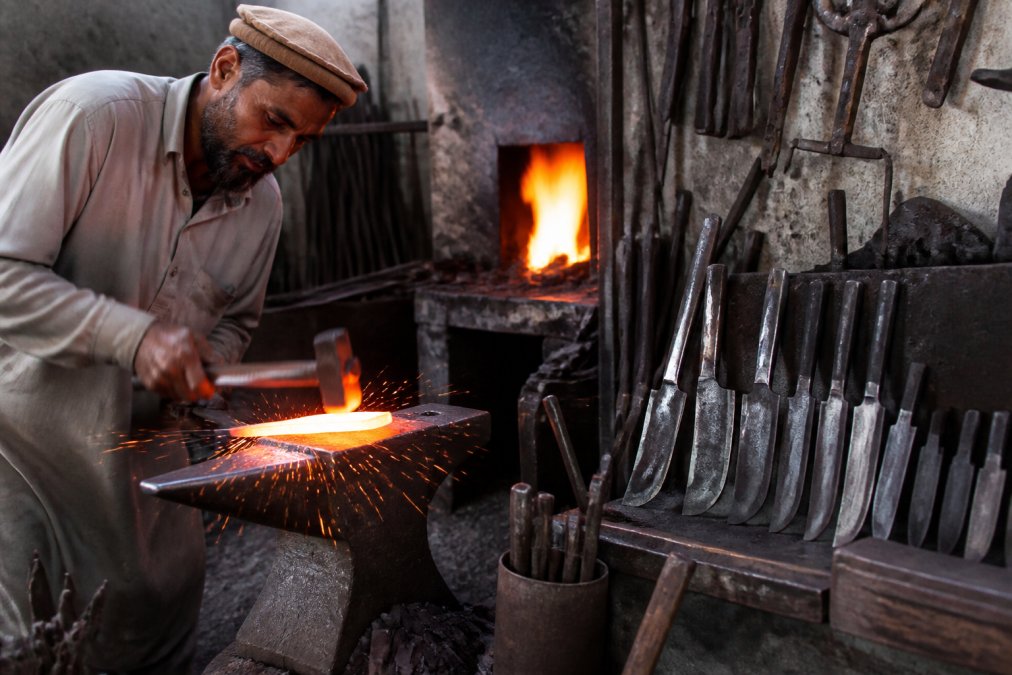 A bladesmith shapes a glowing steel blade on an anvil as sparks fly in a traditional metalworking workshop.