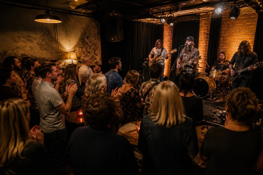 A lively local live music venue with an engaged audience, musicians performing on stage under warm ambient lighting, creating an intimate and joyful community atmosphere