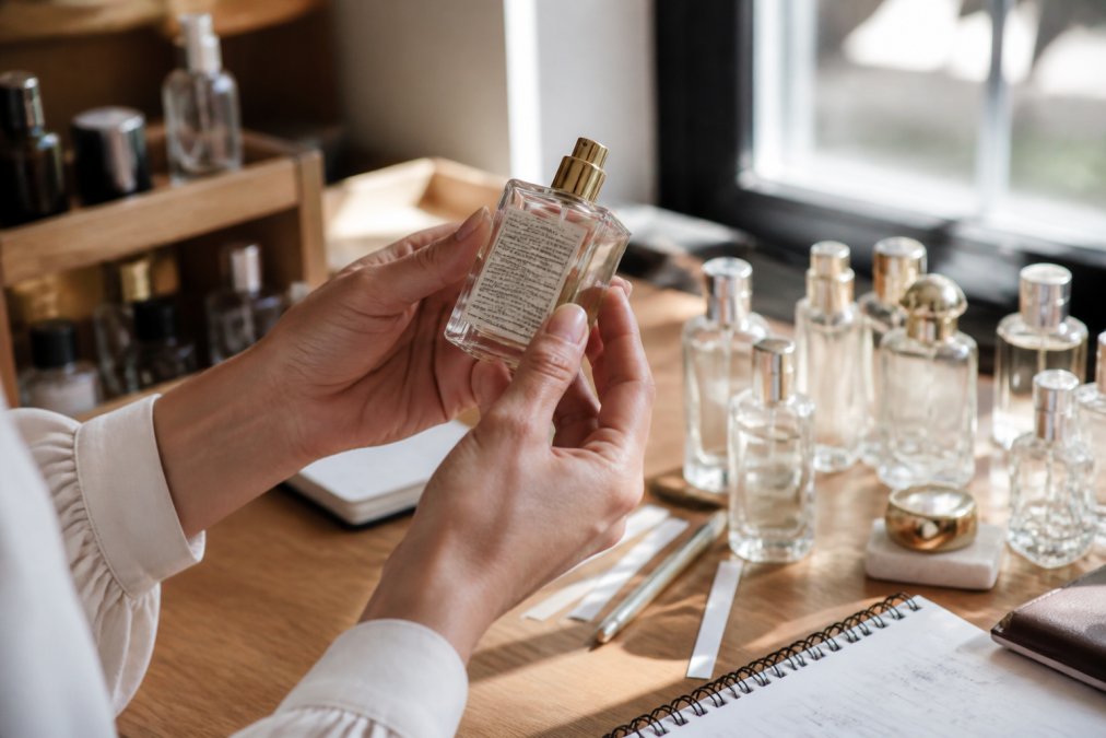 Close-up of hands holding a perfume bottle, carefully examining the ingredient label and back panel text against natural light streaming through a window