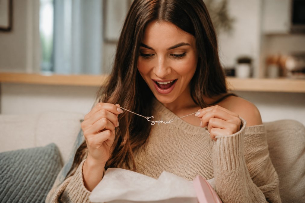 A person opening a wrapped gift with genuine joy, carefully holding a personalized name necklace, their face showing emotional connection and delight