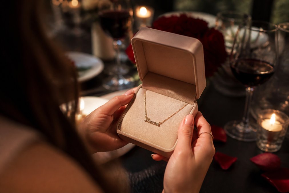 Hands holding a delicate gold name necklace in a gift box, perfect for personalized gifts at a romantic dinner.