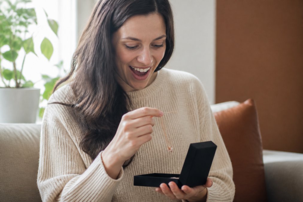 Woman opening a personalized jewelry gift box with genuine emotion, examining a delicate custom necklace with soft natural lighting