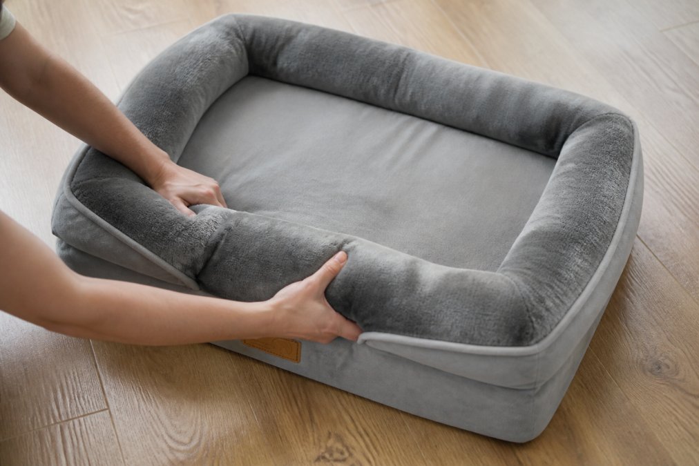 Close-up of hands fluffing and reshaping a compressed dog bed, showing texture being lifted and restored, natural daylight on a wooden floor
