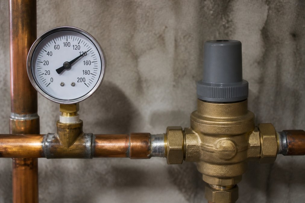 Close-up of a water pressure gauge mounted on copper piping showing high psi reading, with a pressure regulator valve visible nearby in basement lighting