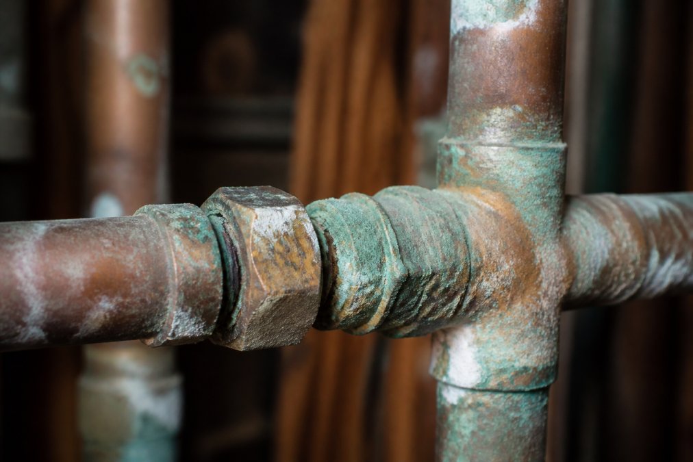 Close-up of corroded copper pipes with green oxidation and damaged compression fittings showing joint failure and mineral deposits in a residential plumbing system under harsh overhead lighting