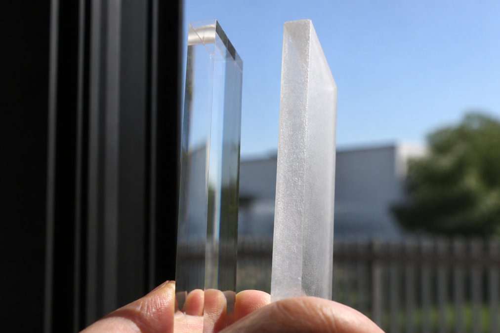 Close-up of a polished acrylic edge held up to bright light, showing crystal-clear transparency and smooth surface finish, contrasted with a rough unpolished edge beside it appearing cloudy and frosted